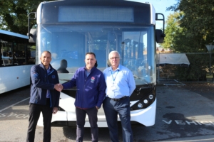 Three men standing in front of a single-deck bus. Two of the men are shaking hands.