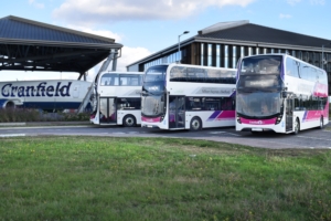 Three double deck buses parked on a field next to an airplane at Cranfield.