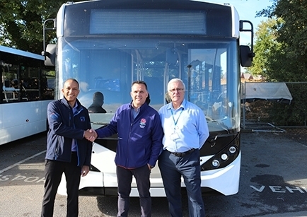 Three men standing in front of a white single-deck bus. Two men are shaking hands.