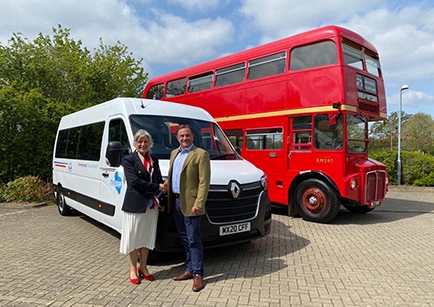 Two people shaking hands in front of a white minibus and a Routemaster