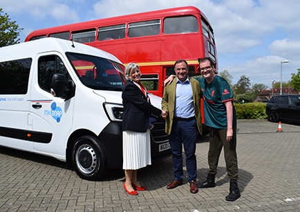 Three people standing in front of a minibus and double-deck bus. Two are shaking hands.